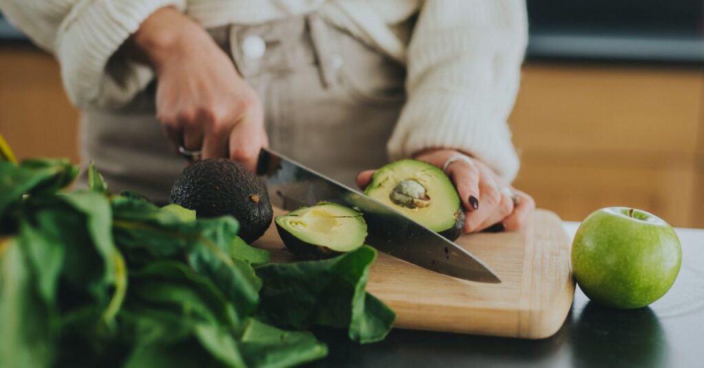 A woman slicing an avocado next to a plate of leafy greens