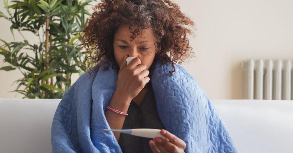 A woman blowing her nose and reading her temperature off a digital thermometer
